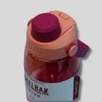 A translucent red water bottle with a peach-colored lid and a dark magenta flip-top cap, featuring a white label with the brand name 'KELBAK'.